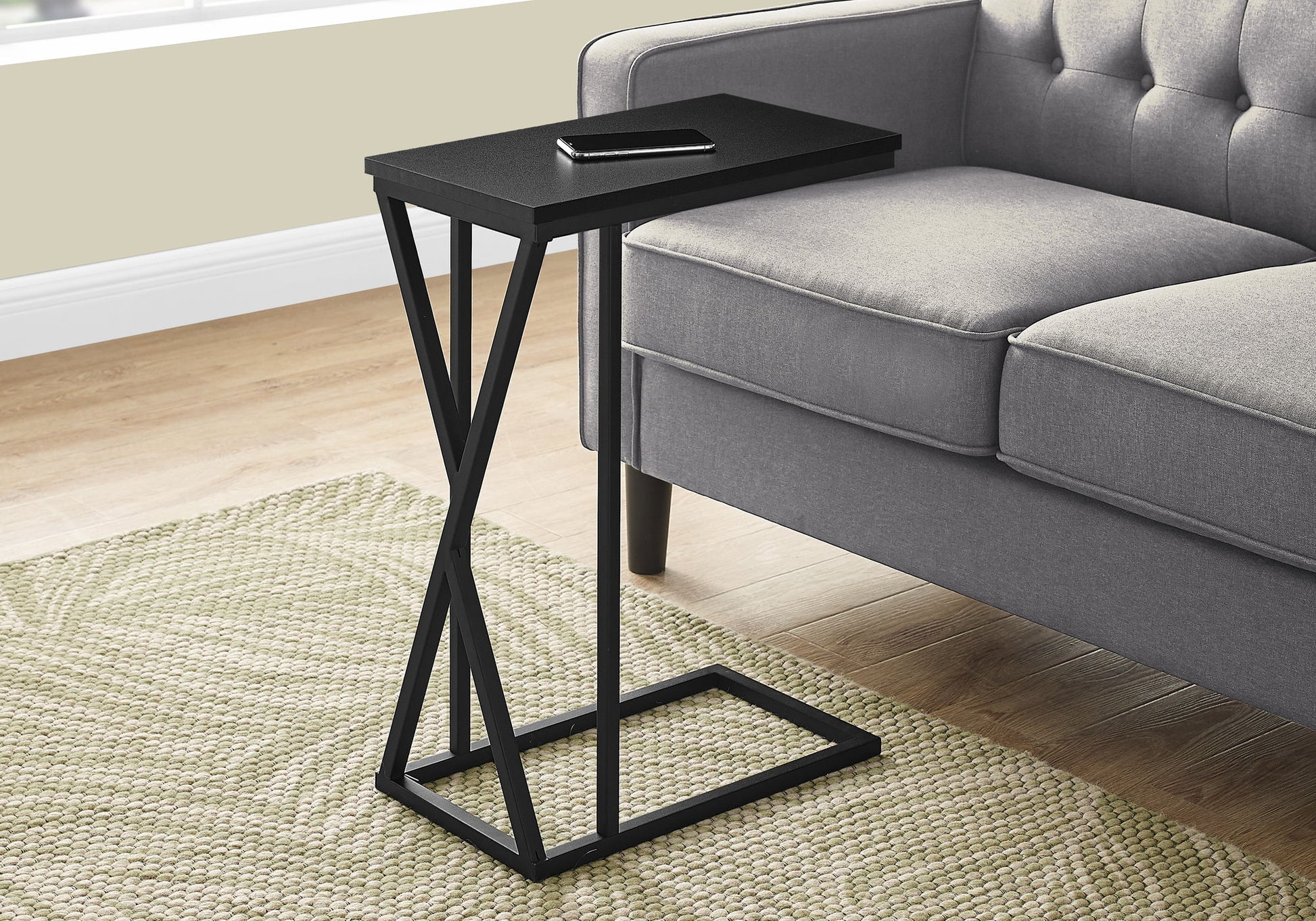 The Accent Table C - Shaped, Contemporary & Modern stands by a gray sofa on a light green textured rug. A smartphone rests atop the small black table with a geometric metal frame; wood floors and a light wall complete the setting.