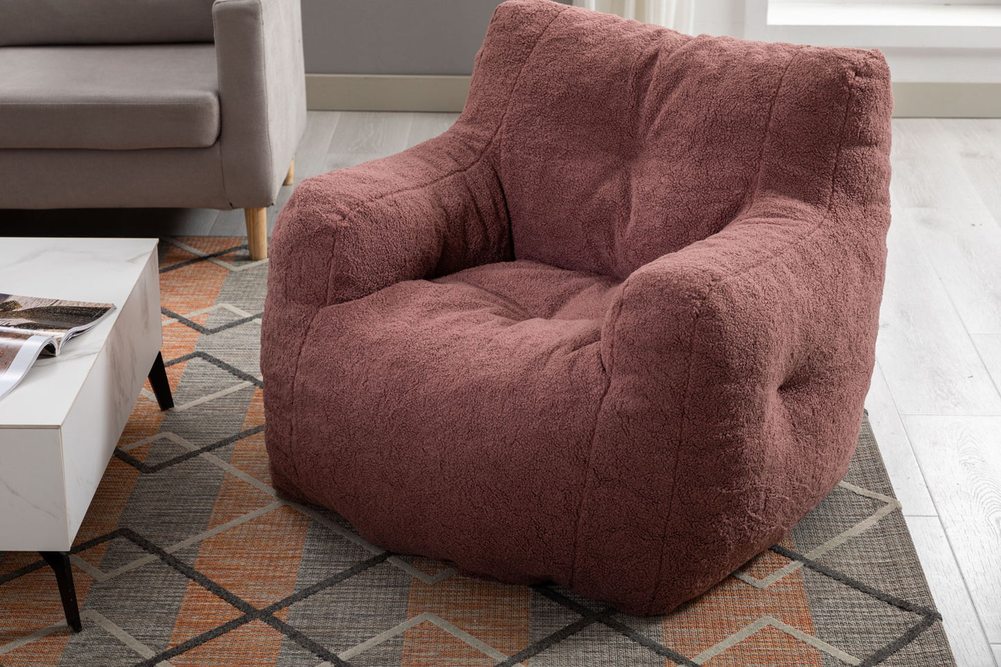 A Soft Teddy Fabric Tufted Foam Bean Bag Chair sits on a patterned rug beside a white coffee table and beige sofa in a modern living room with light wood flooring.