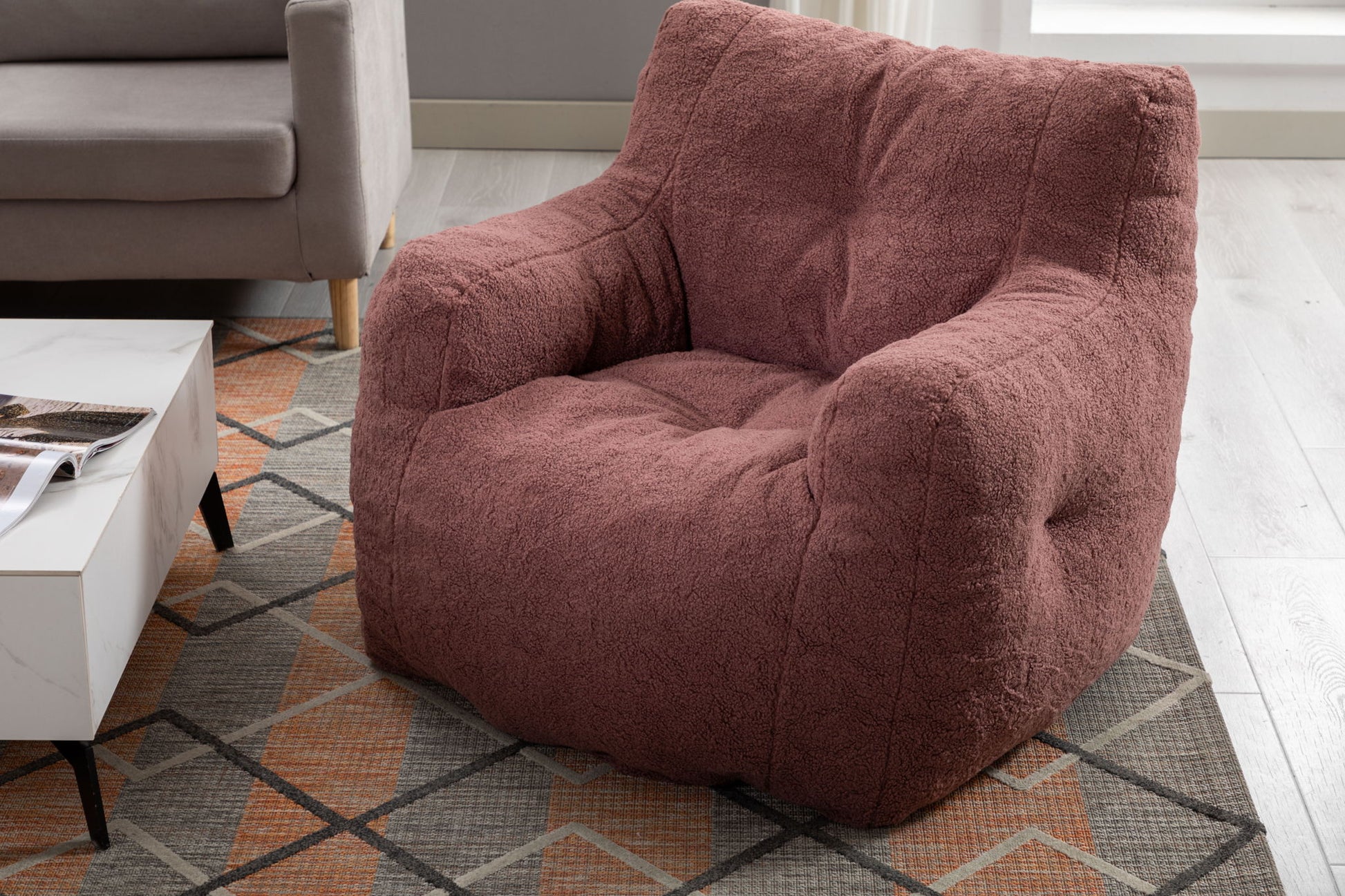 A Soft Teddy Fabric Tufted Foam Bean Bag Chair sits on a patterned rug beside a white coffee table and beige sofa in a modern living room with light wood flooring.