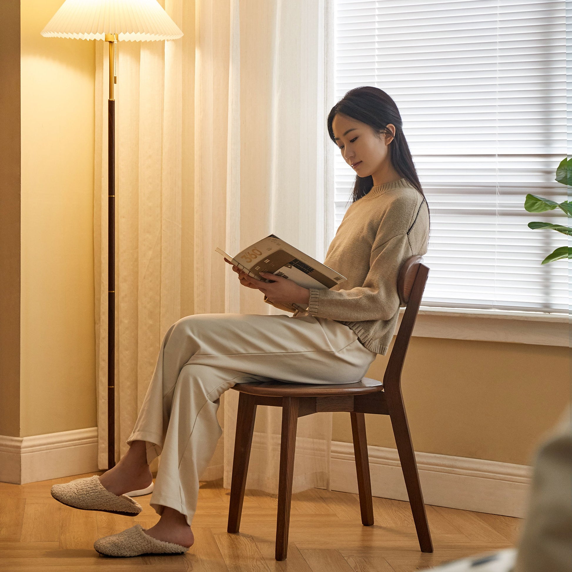 A woman sits on a Simple Dining Chair, Solid Chair Table For Living Room Chair in a cozy room, reading a book. She wears a beige sweater, white pants, and slippers. A lamp and window with blinds add to the tranquil ambiance.