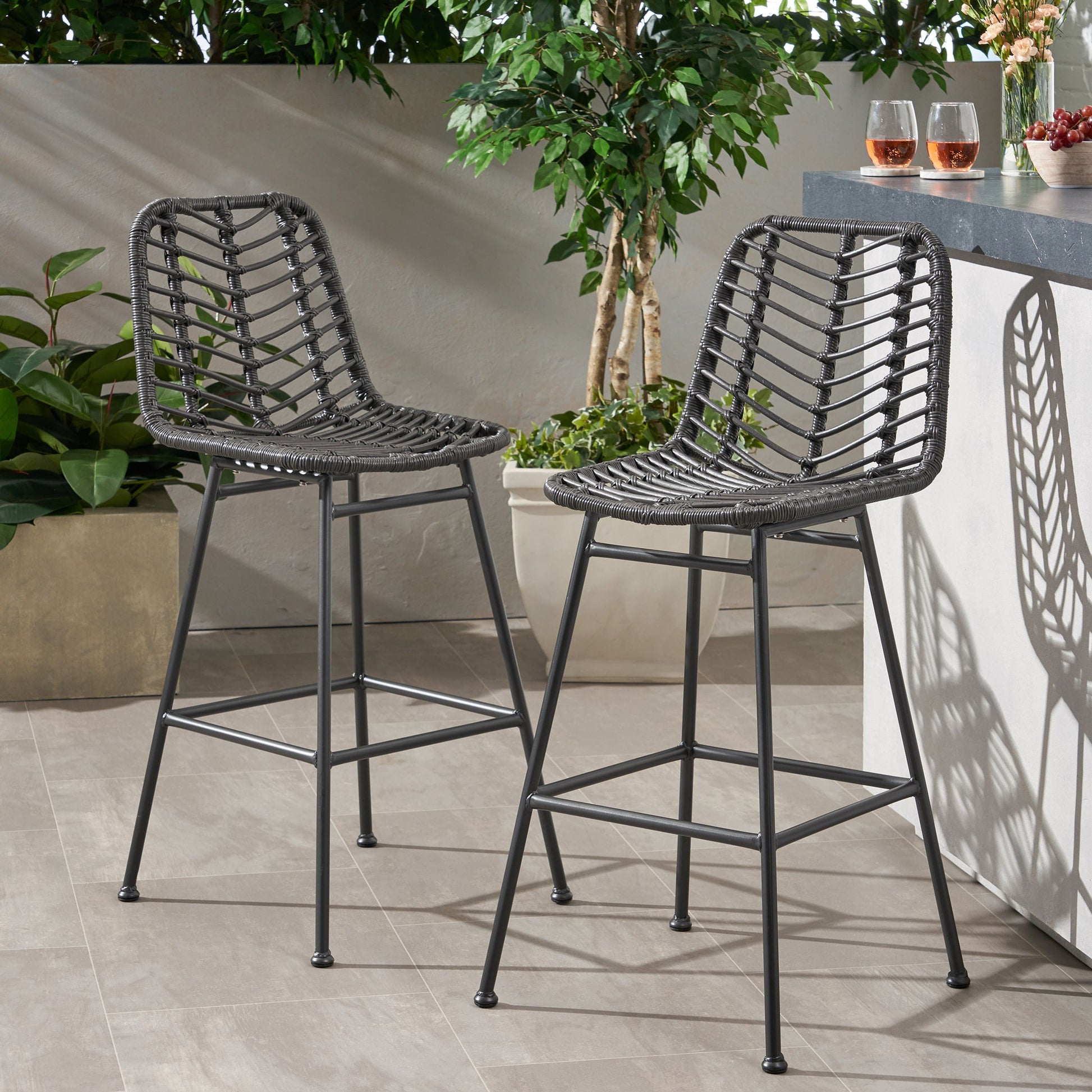 Two Sawtelle Outdoor Wicker Barstools (Set of 2) with black wicker seats and metal legs sit on a tiled patio beside a bar counter. Green plants and three drinks in glasses are seen in the background, with sunlight casting floor shadows.