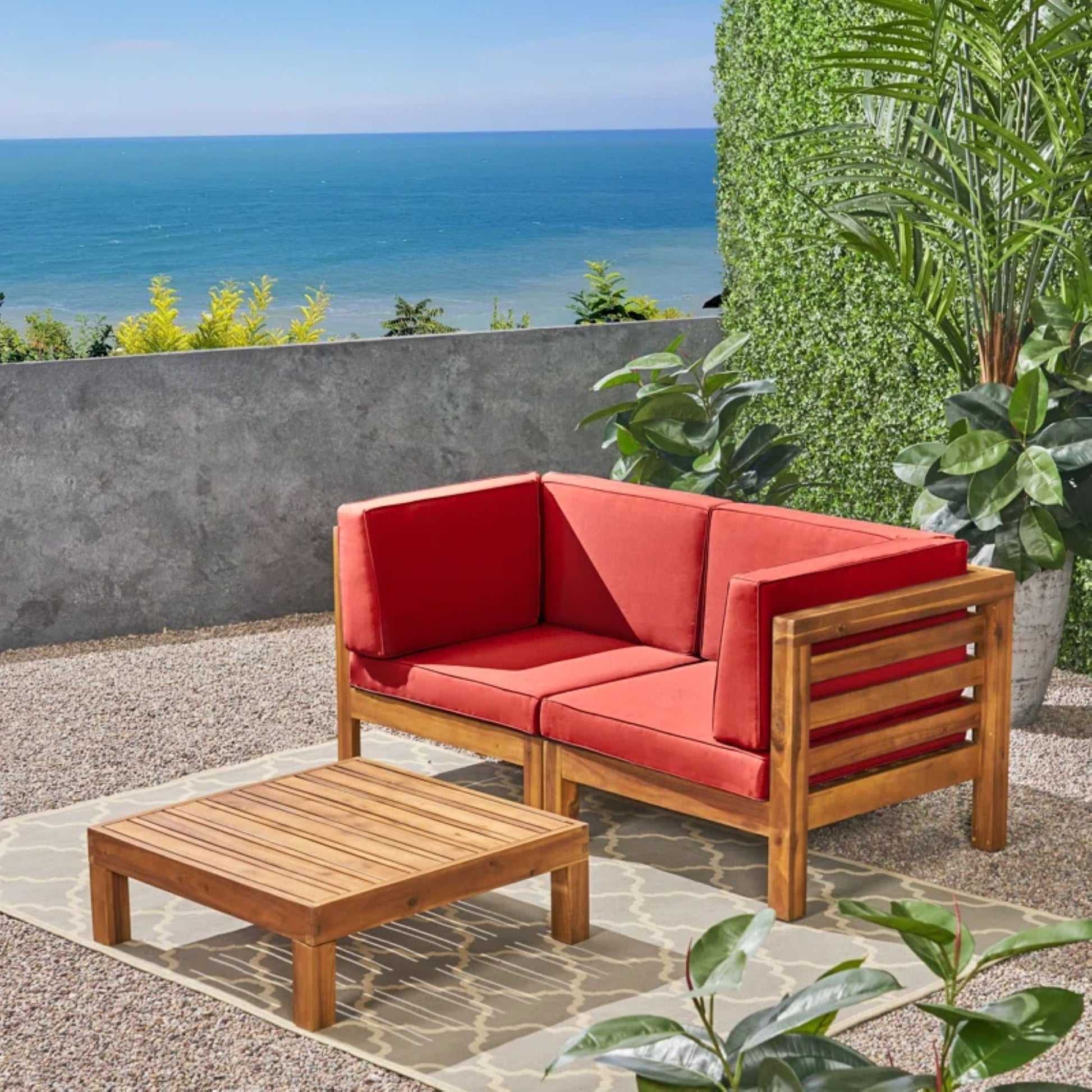 The Oana 3 Piece Love Seat Set with Coffee Table, featuring red cushions, rests on a patio rug amid lush greenery, offering an ocean view under a clear blue sky.