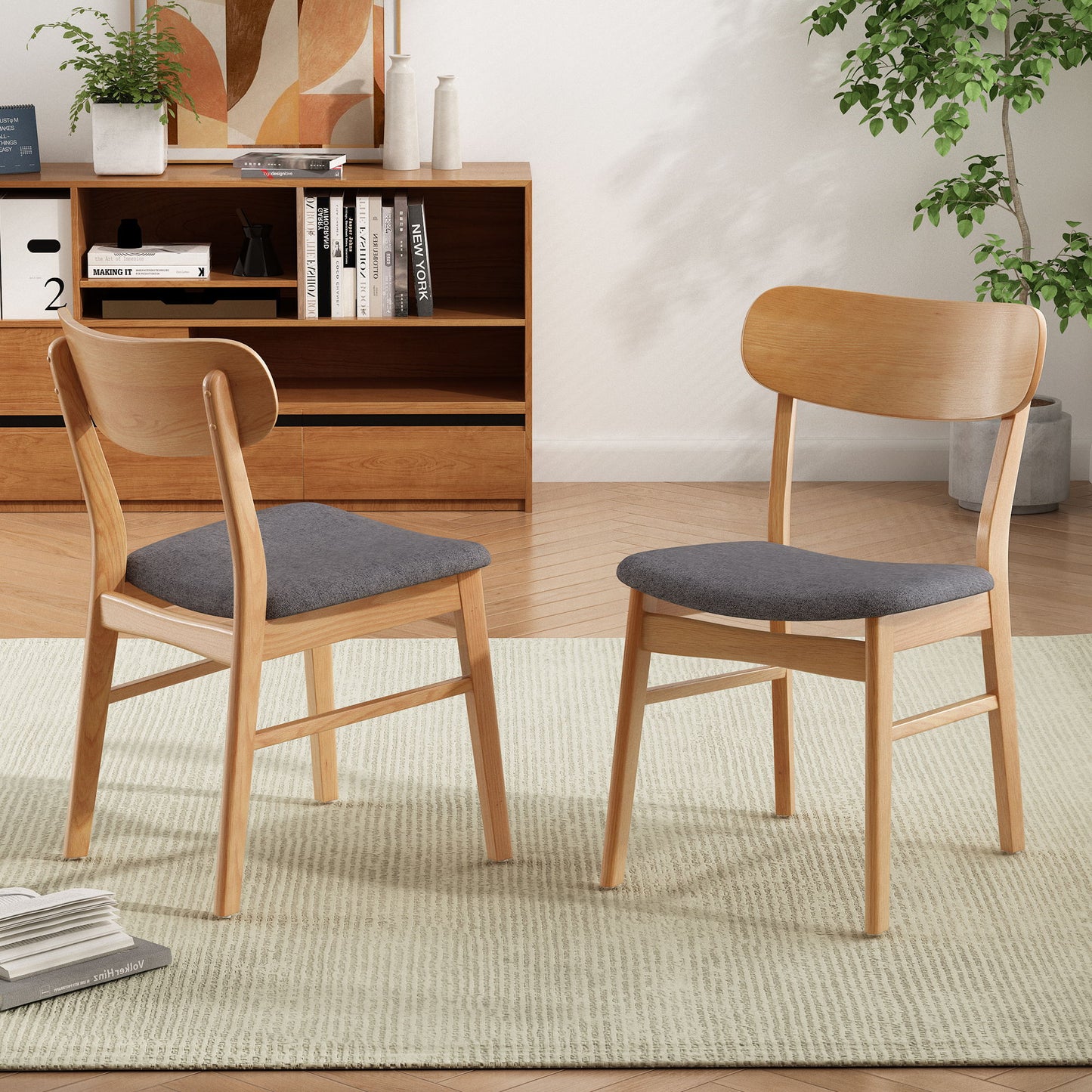 A set of two Brilliantine Dining Chairs with gray cushions and wood frames sit on a light rug in a modern living room, complemented by a wooden shelf, books, decor, and a leafy plant in the background.