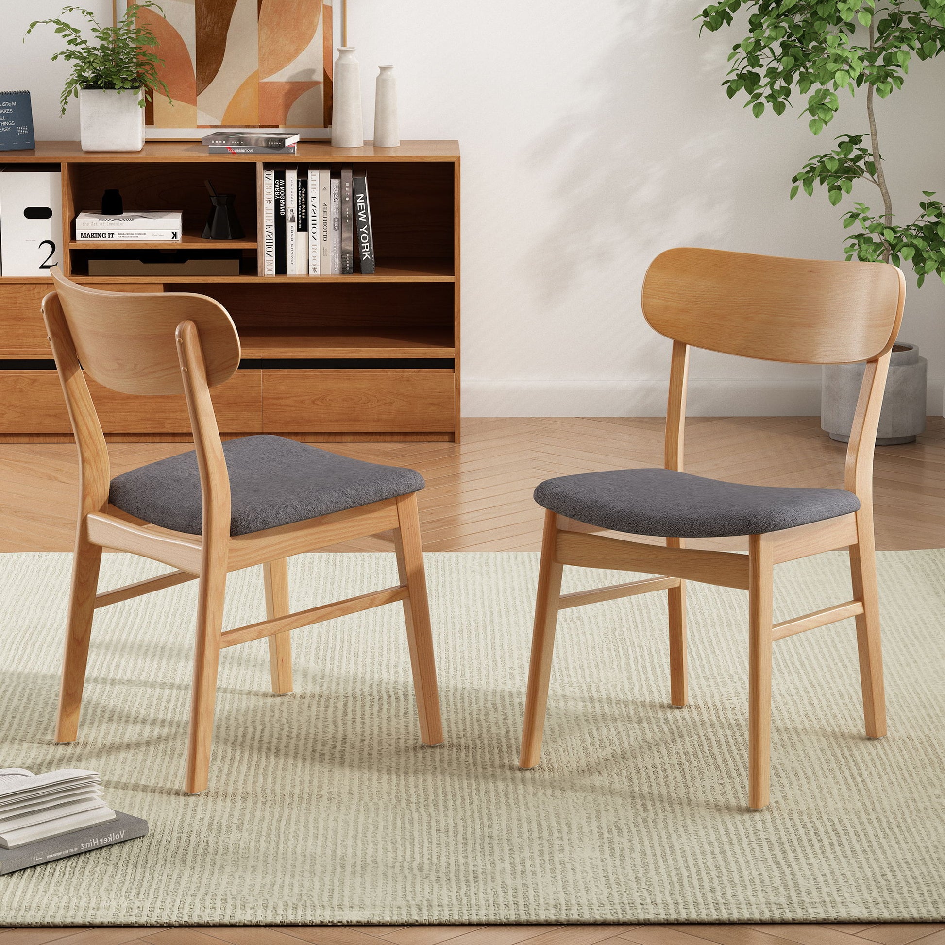 A set of two Brilliantine Dining Chairs with gray cushions and wood frames sit on a light rug in a modern living room, complemented by a wooden shelf, books, decor, and a leafy plant in the background.