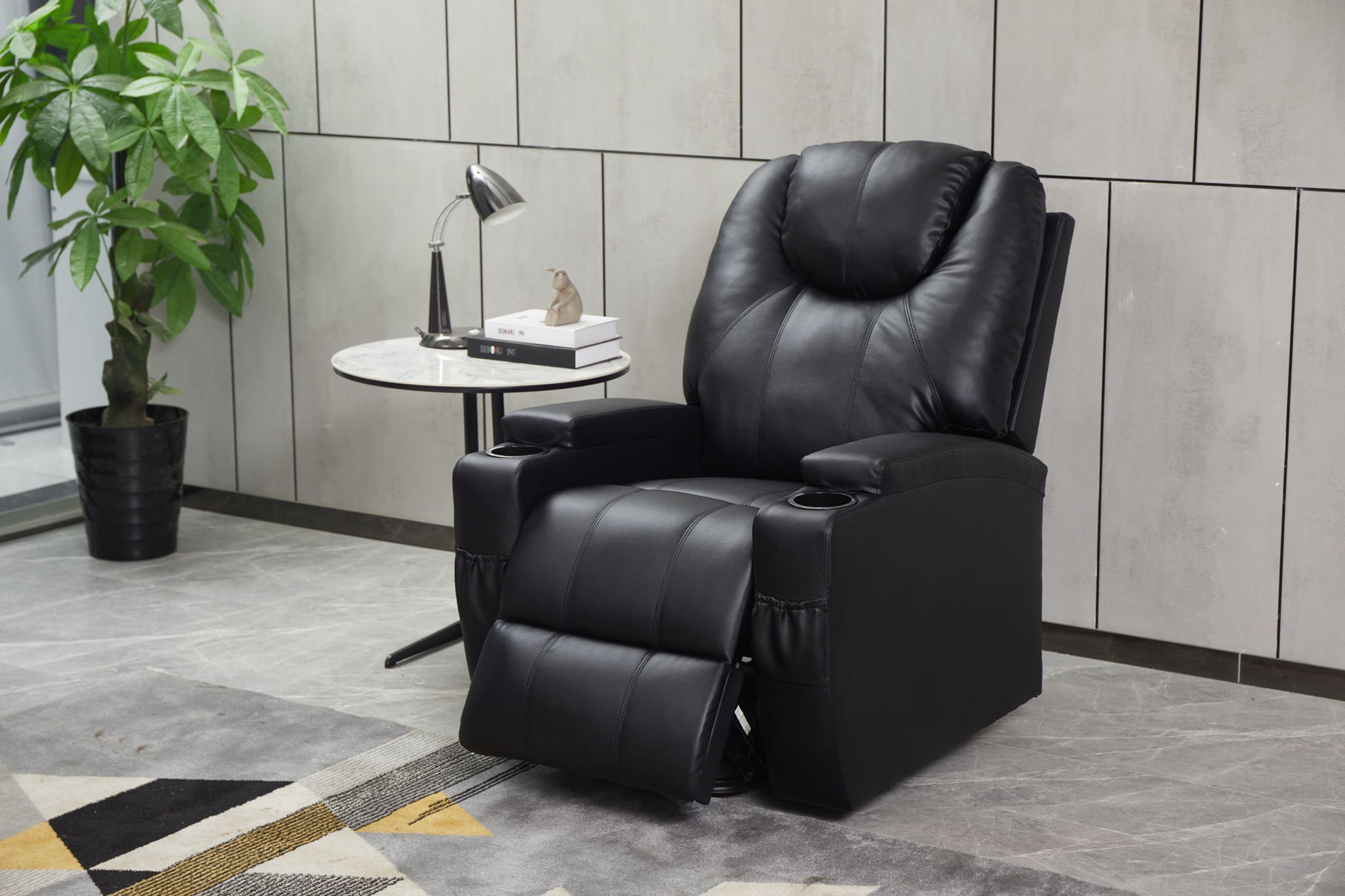 A Modern Recliner Chair With Cup Holders and Remote Control, footrest extended, sits beside a round table with a lamp, books, and sculpture. A potted plant is nearby against the tiled wall.