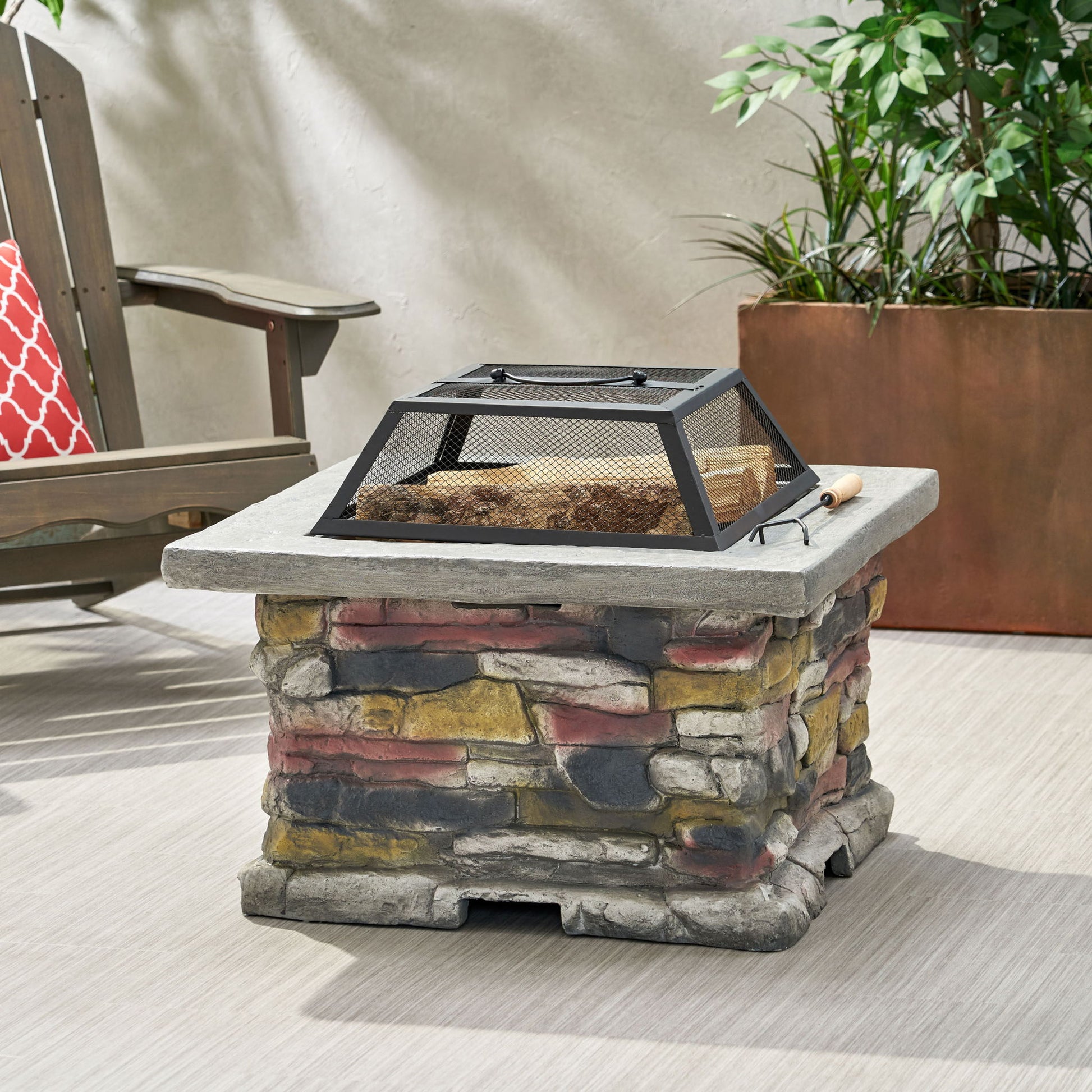 An Outdoor Wood Burning Magnesium Oxide Fire Pit with a lid features a stone-patterned base and mesh cover, situated on a patio near a potted plant and wooden chair with a red patterned pillow.