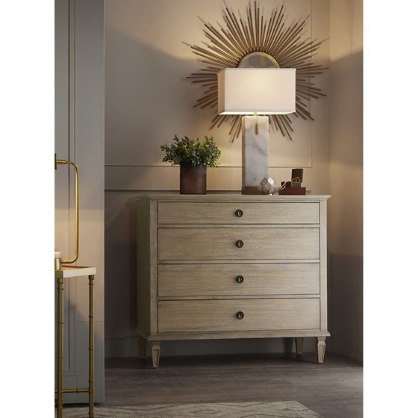 The French Inspired Small Dresser - Wire Brushed Light Natural stands against the wall, topped with a potted plant, decor, and a marble lamp. Above it, a gold sunburst mirror adds warmth and elegance to the room.