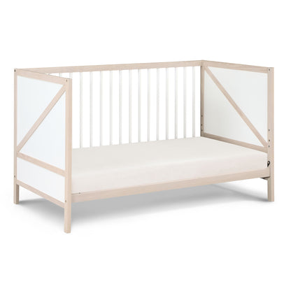 The Pixie Zen 3-in-1 Crib in walnut features a light wood frame, white geometric panel sides, white vertical slats on the back, and includes a plain white mattress.