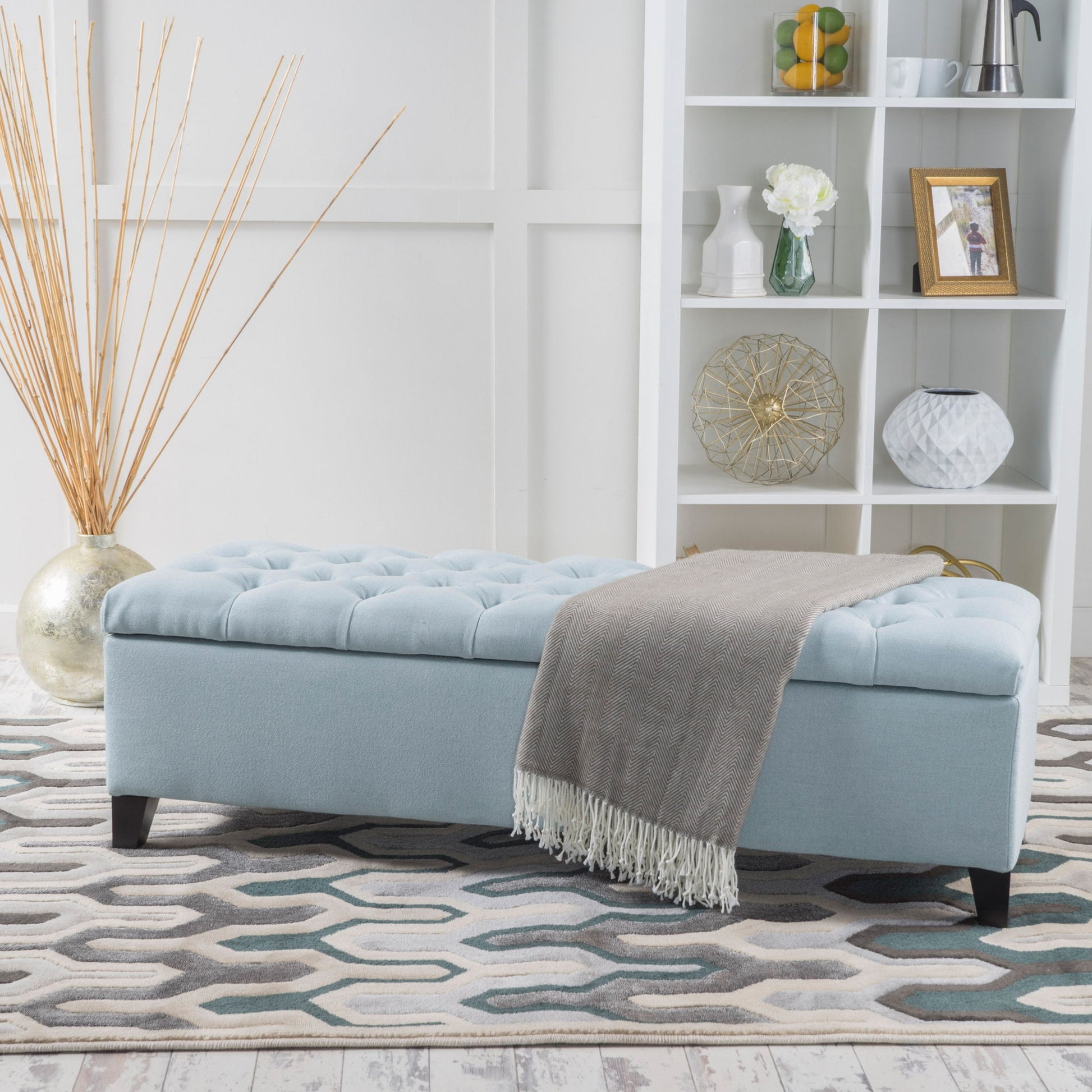 The Glouster Contemporary Storage Ottoman with button tufted design in light blue sits on a patterned rug, draped with a gray throw. Behind it stands a white shelving unit displaying decor and a tall vase filled with reed sticks.
