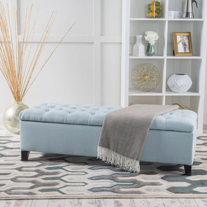 The Glouster Contemporary Storage Ottoman with button tufted design in light blue sits on a patterned rug, draped with a gray throw. Behind it stands a white shelving unit displaying decor and a tall vase filled with reed sticks.