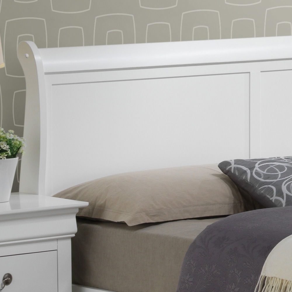 A close-up of the Louis Phillipe - Storage Bed in white wood, styled with a beige pillow and gray patterned blanket, next to a matching white nightstand holding a potted plant, all set against a gray geometric patterned wall.