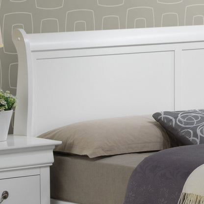 A close-up of the Louis Phillipe - Storage Bed in white wood, styled with a beige pillow and gray patterned blanket, next to a matching white nightstand holding a potted plant, all set against a gray geometric patterned wall.