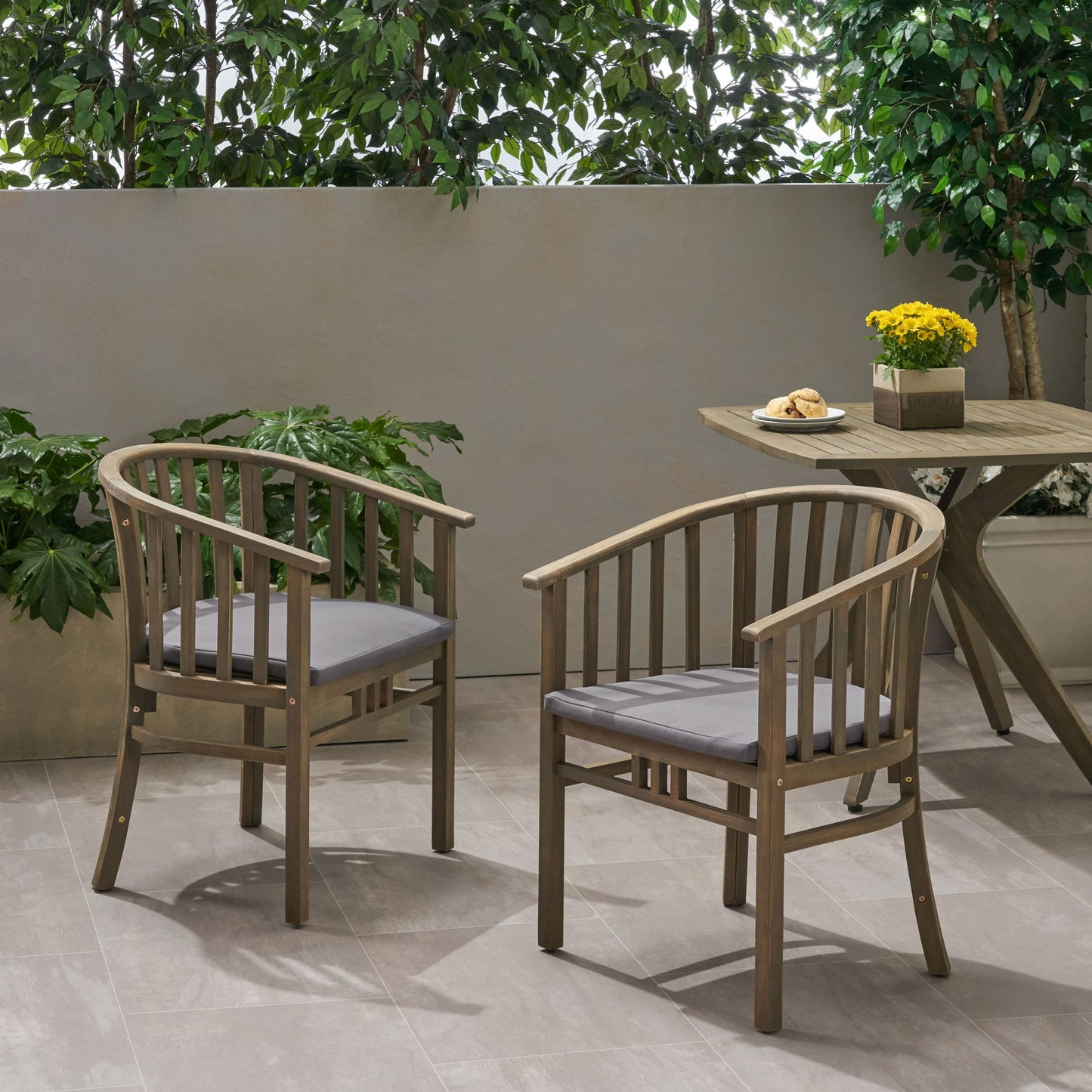Two Outdoor Wooden Dining Chairs With Cushions (Set of 2) sit on a tiled patio next to a round table with yellow flowers and pastries, surrounded by potted greenery and a leafy backdrop.
