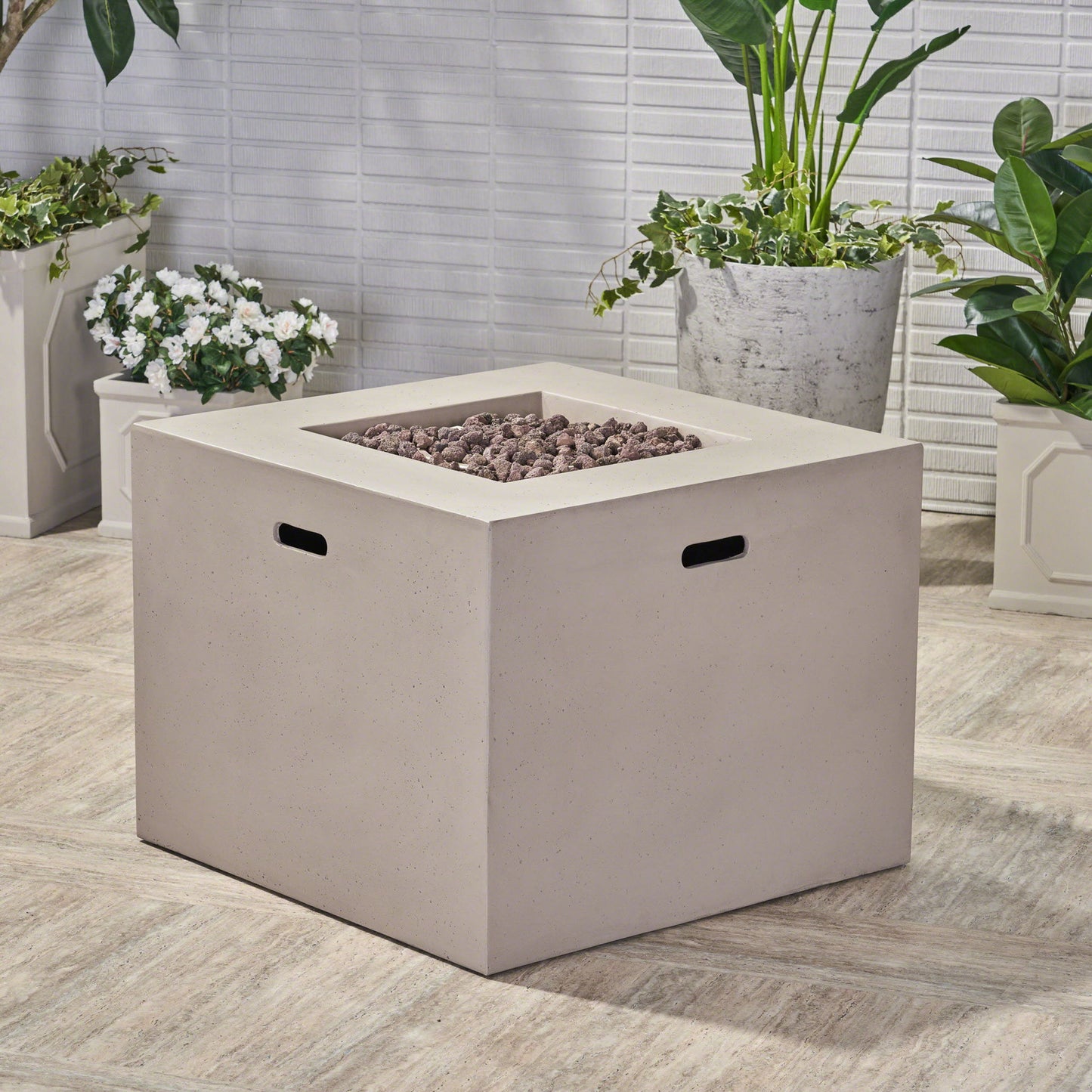 The Outdoor Patio Square Gas Burning Concrete Fire Pit 40,000 Btu, featuring black handles and a beige finish, is set on a tiled patio surrounded by potted green plants and white flowers against a white brick wall.