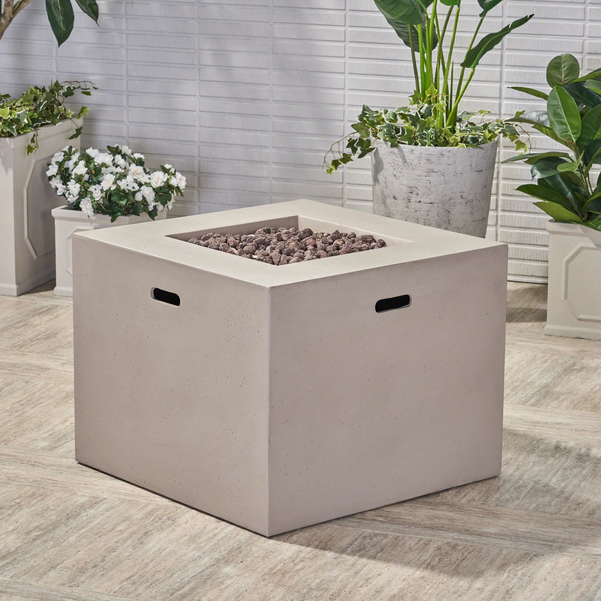 The Outdoor Patio Square Gas Burning Concrete Fire Pit 40,000 Btu, featuring black handles and a beige finish, is set on a tiled patio surrounded by potted green plants and white flowers against a white brick wall.