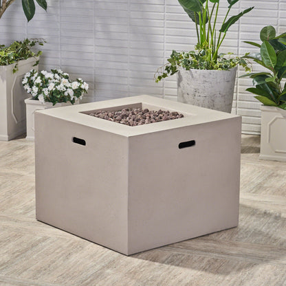 The Outdoor Patio Square Gas Burning Concrete Fire Pit 40,000 Btu, featuring black handles and a beige finish, is set on a tiled patio surrounded by potted green plants and white flowers against a white brick wall.