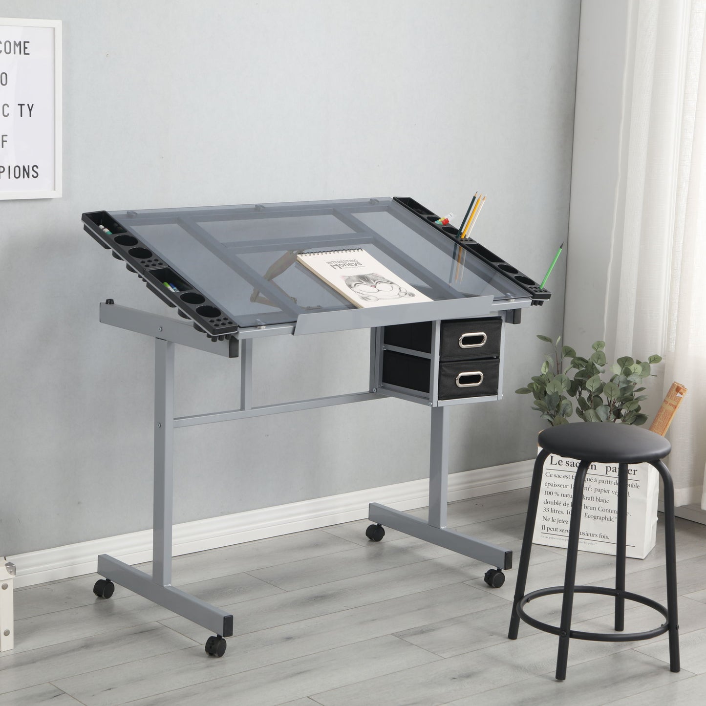 An Adjustable Art Drawing Desk Craft Station Drafting with a glass top, angled surface, 2 non-woven fabric slide drawers, and 4 wheels—shown with art supplies and a black stool in a bright room with light wood floors and tall windows.