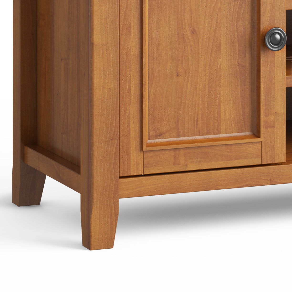 Close-up of the Amherst - TV Media Stand, Handcrafted, showing its lower corner with a rectangular door, metal round handle, tapered legs, and warm medium-brown wood finish with visible grain.