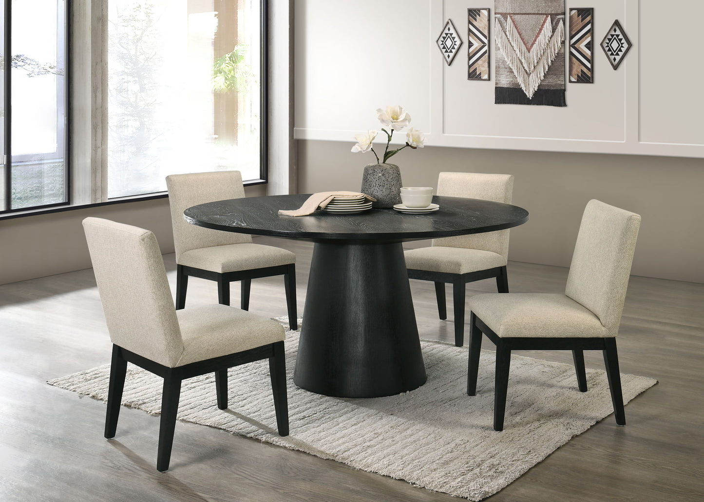 A modern dining room featuring the Froja Manufactured Wood Round Dining Table in black, four beige upholstered chairs, a gray rug, and a decorative flower vase. Large windows and wall art appear in the background.