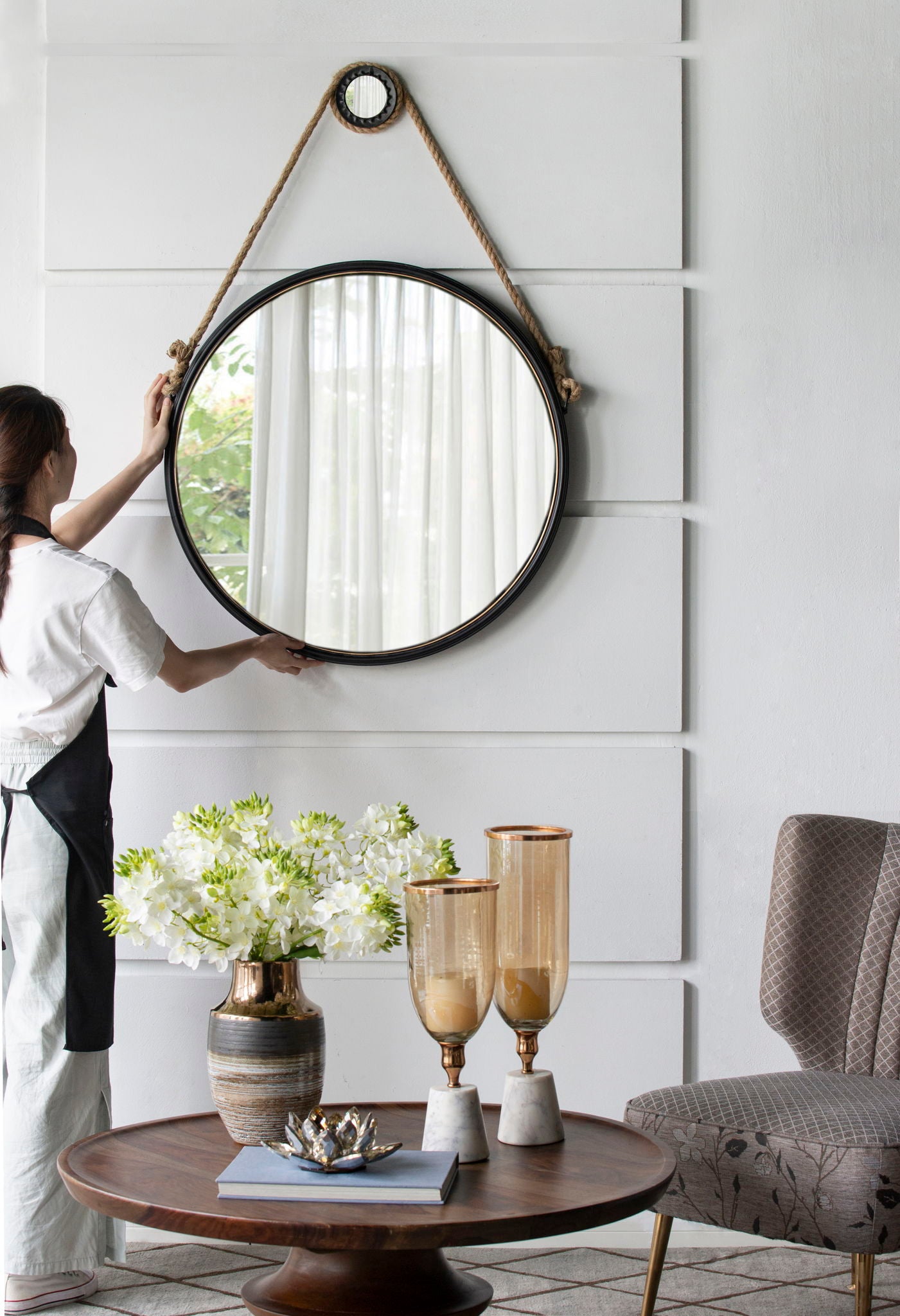 A woman adjusts the Handsome Cleveland Mirror With Rope Strap, a contemporary round metal-framed mirror, on a white wall. Below, a table has a vase of white flowers, amber glass candle holders, and a book. A gray chair is nearby; light filters through sheer curtains.