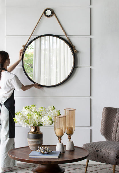 A woman adjusts the Handsome Cleveland Mirror With Rope Strap, a contemporary round metal-framed mirror, on a white wall. Below, a table has a vase of white flowers, amber glass candle holders, and a book. A gray chair is nearby; light filters through sheer curtains.