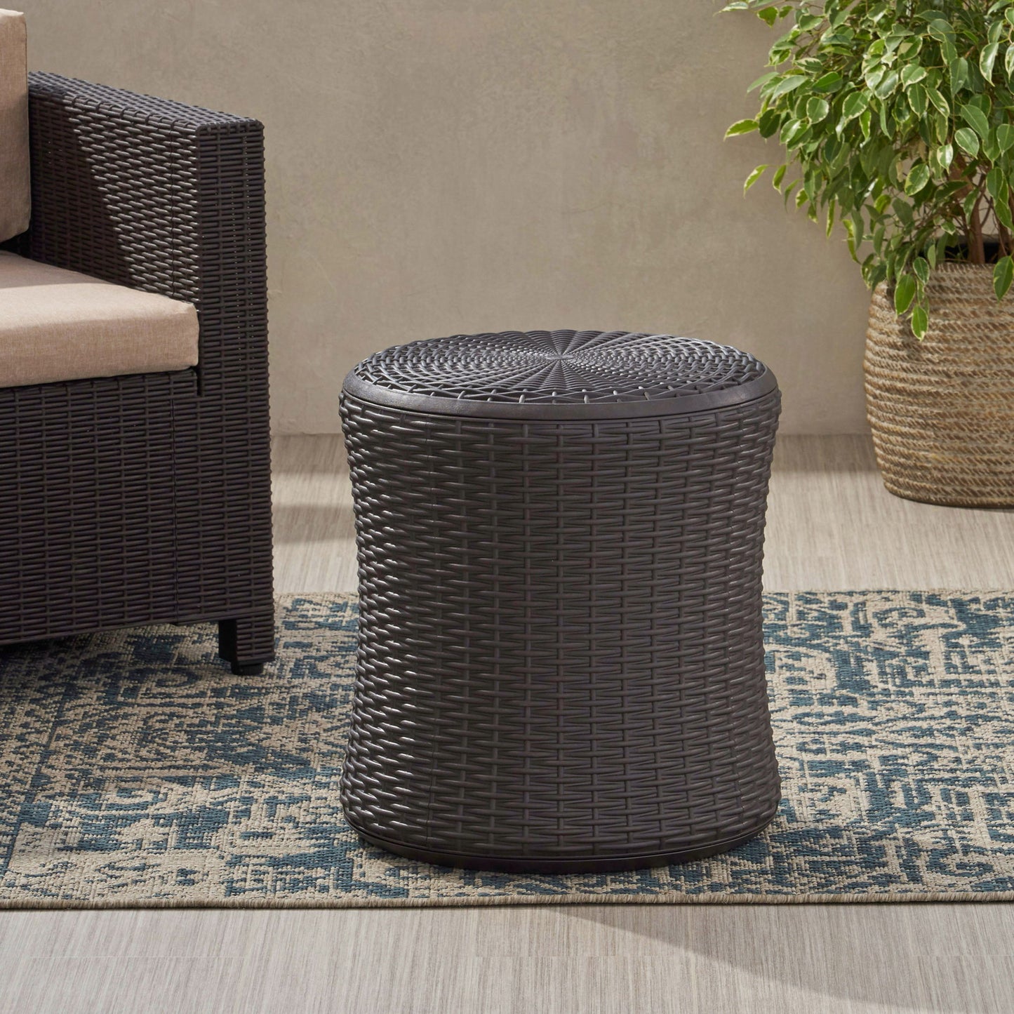 The Olivo Patio Table Outdoor Side Table in dark brown sits on a blue patterned rug beside a wicker armchair and leafy potted plant in a light, wood-floored room.