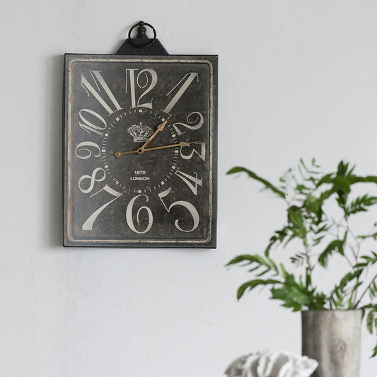 The Large Vintage Rectangular Wall Clock With Numerals in white and black, featuring two gold hands, hangs on a light gray wall, with a green potted plant visible in the foreground on the right.