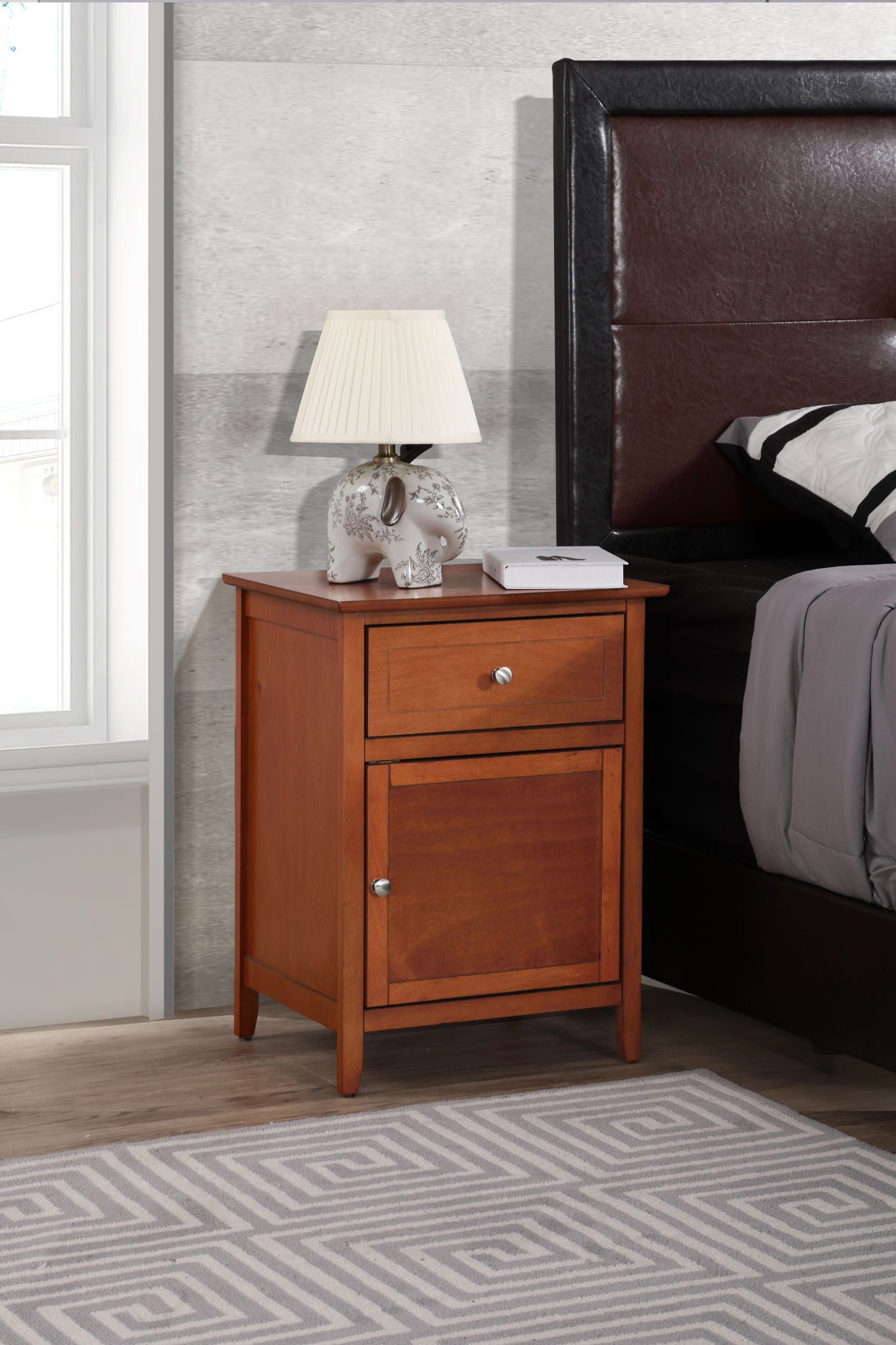 The Izzy 1 Drawer / 1 Door Nightstand stands beside a bed with a brown leather headboard. An elephant-shaped lamp with a white shade rests on the nightstand, while a geometric rug and window complete the scene.