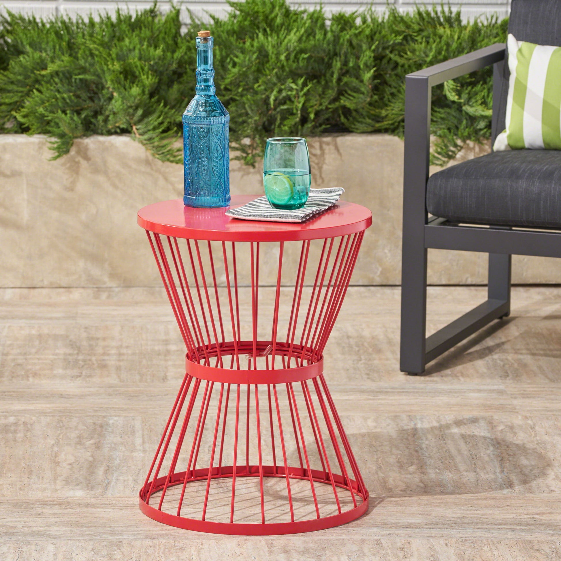 A Lassen Modern Outdoor Side Table with an hourglass design holds a bottle, glass, and striped napkins on a patio near a gray chair with a green-striped pillow, set against greenery.