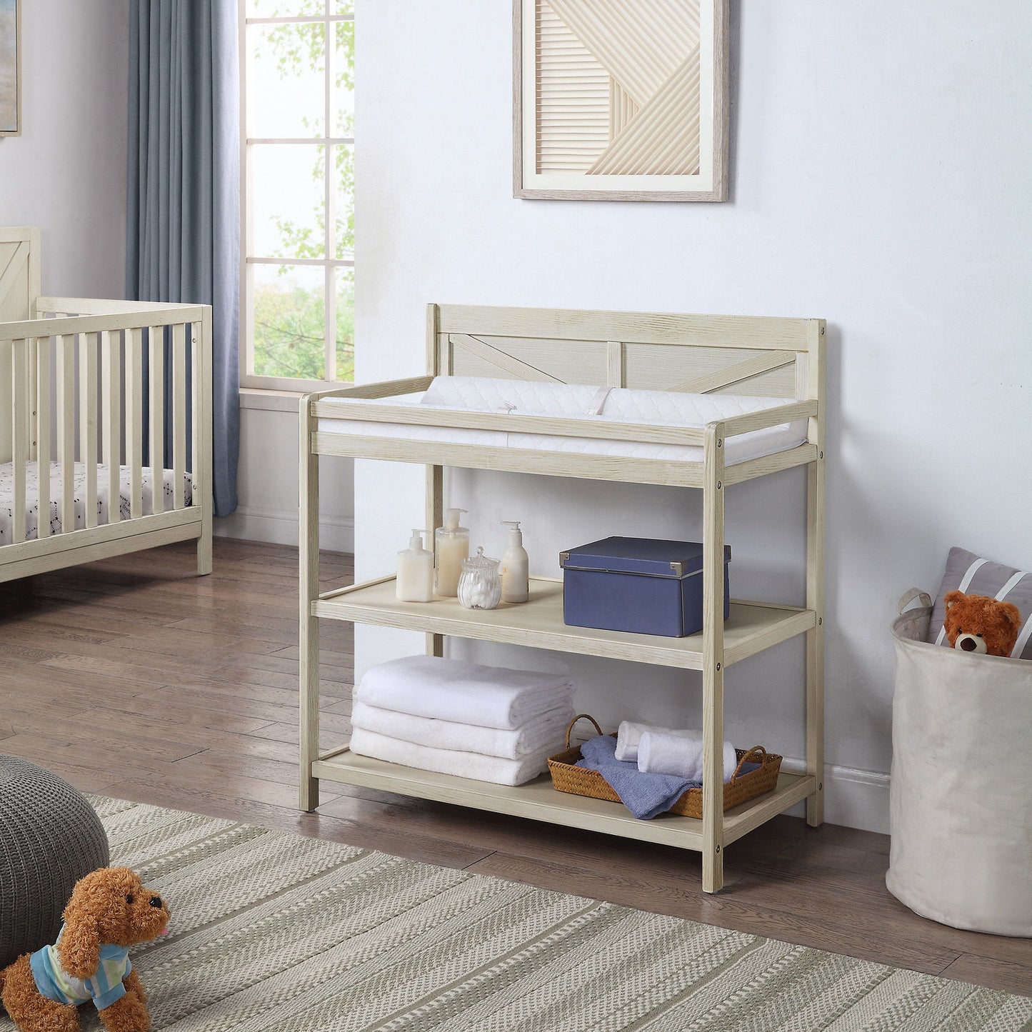 The Barnside - Changing Table Vintage features a cushioned pad, open shelves for towels, bottles, and storage. Behind it in the nursery are a crib, window, and a basket holding a teddy bear.