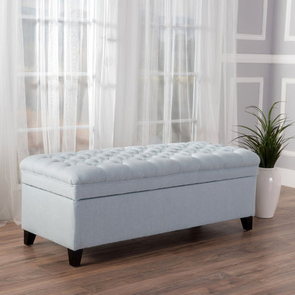 The Juliana Storage Ottoman with birch legs, in light blue tufted fabric, stands on a wooden floor before tall windows with sheer curtains. Nearby, a potted plant rests against a gray paneled wall.