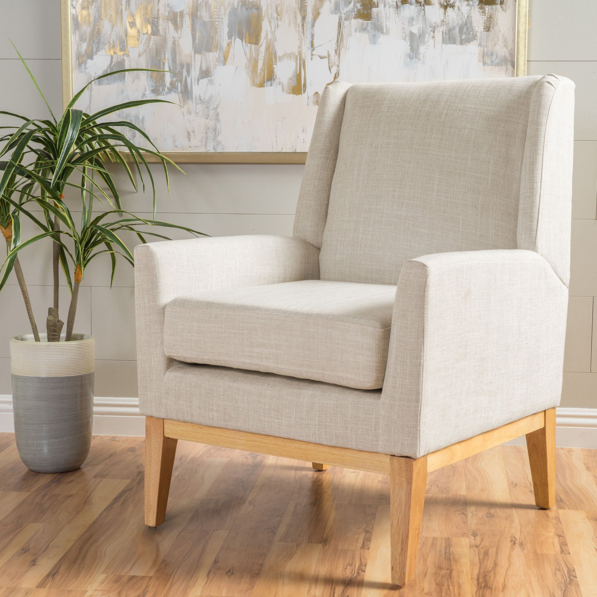 The Modern Accent Chair With Padded Cushion Seat features light beige upholstery and wooden legs on a hardwood floor, paired with a potted plant nearby and an abstract painting on the light gray wall behind it.