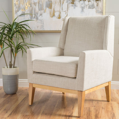 The Modern Accent Chair With Padded Cushion Seat features light beige upholstery and wooden legs on a hardwood floor, paired with a potted plant nearby and an abstract painting on the light gray wall behind it.