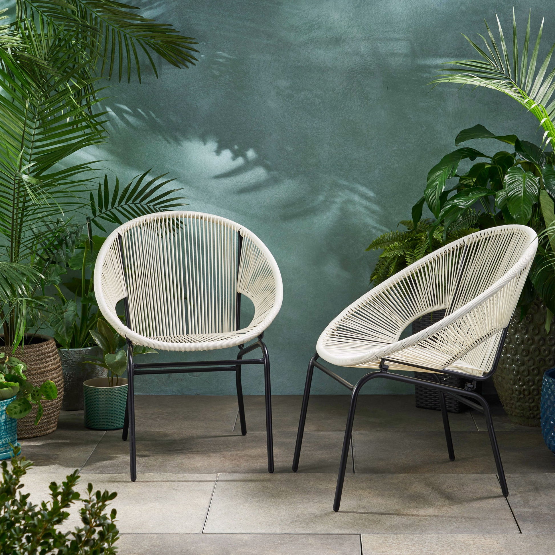 Two Nusa - Modern Chair Outdoor Seating pieces, featuring white round seats and black metal legs, are arranged on a tiled patio with lush green potted plants around them and a textured green wall in the background.