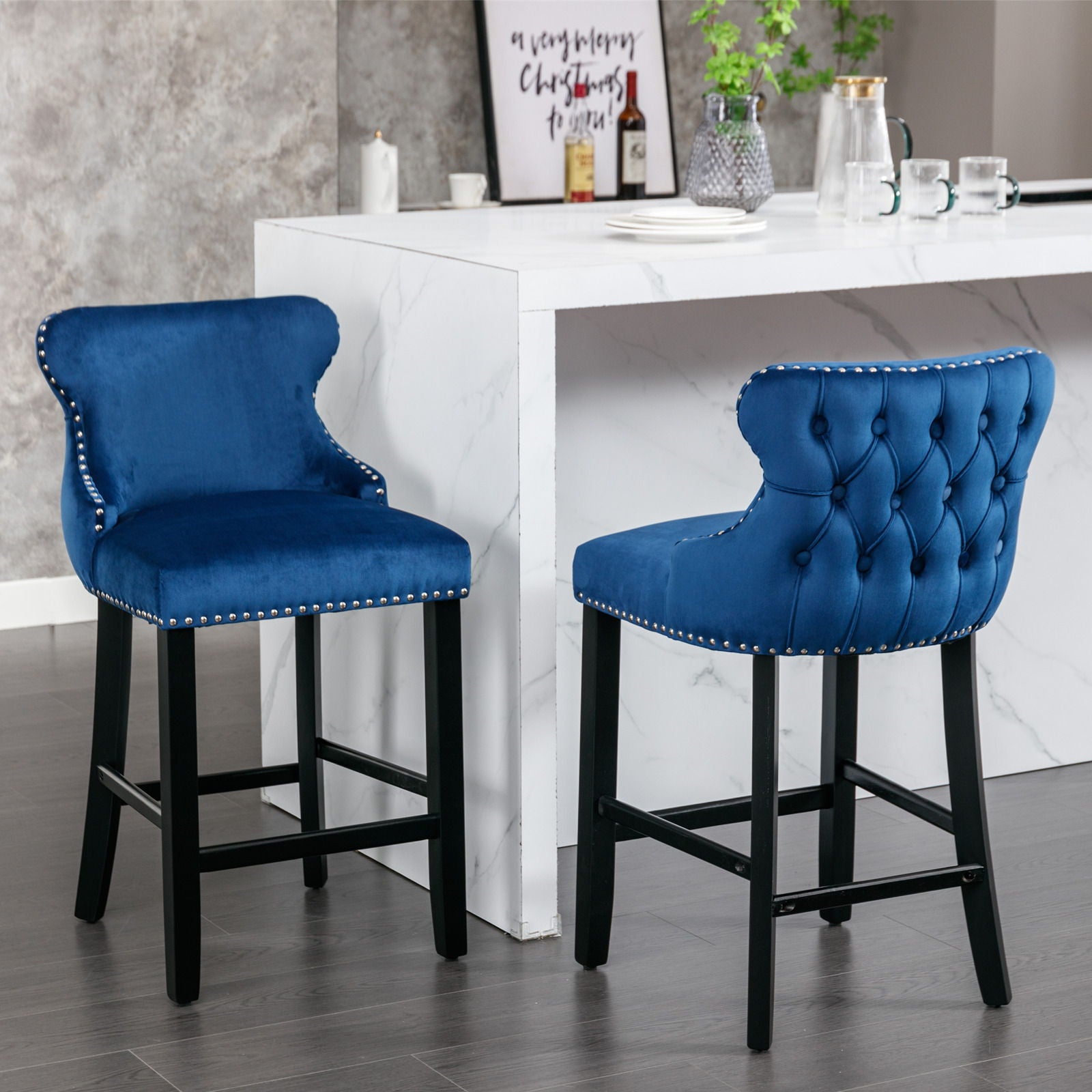 Two Contemporary Velvet Upholstered Wing-Back Barstools with button tufted decoration, wooden legs, and chrome nailhead trim sit in front of a white marble kitchen island topped with cups, a teapot, and decorative accents.
