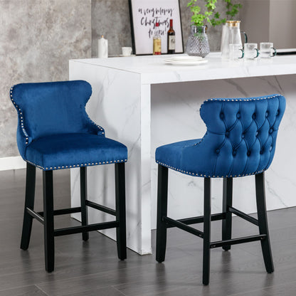 Two Contemporary Velvet Upholstered Wing-Back Barstools with button tufted decoration, wooden legs, and chrome nailhead trim sit in front of a white marble kitchen island topped with cups, a teapot, and decorative accents.
