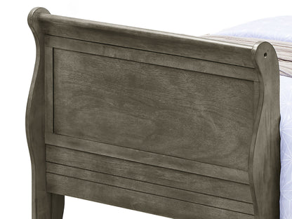 A close-up of the Panel Sleigh Bed’s wooden footboard in dark gray shows its panel design with curved top edges and visible wood grain. A portion of a mattress and blanket is seen in the background.
