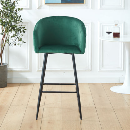 A Counter Height Bar Stool with green velvet upholstery and black metal legs (Set of 2) stands on a light wood floor next to a small white table with red wine, against a backdrop of a potted plant and white paneled wall.