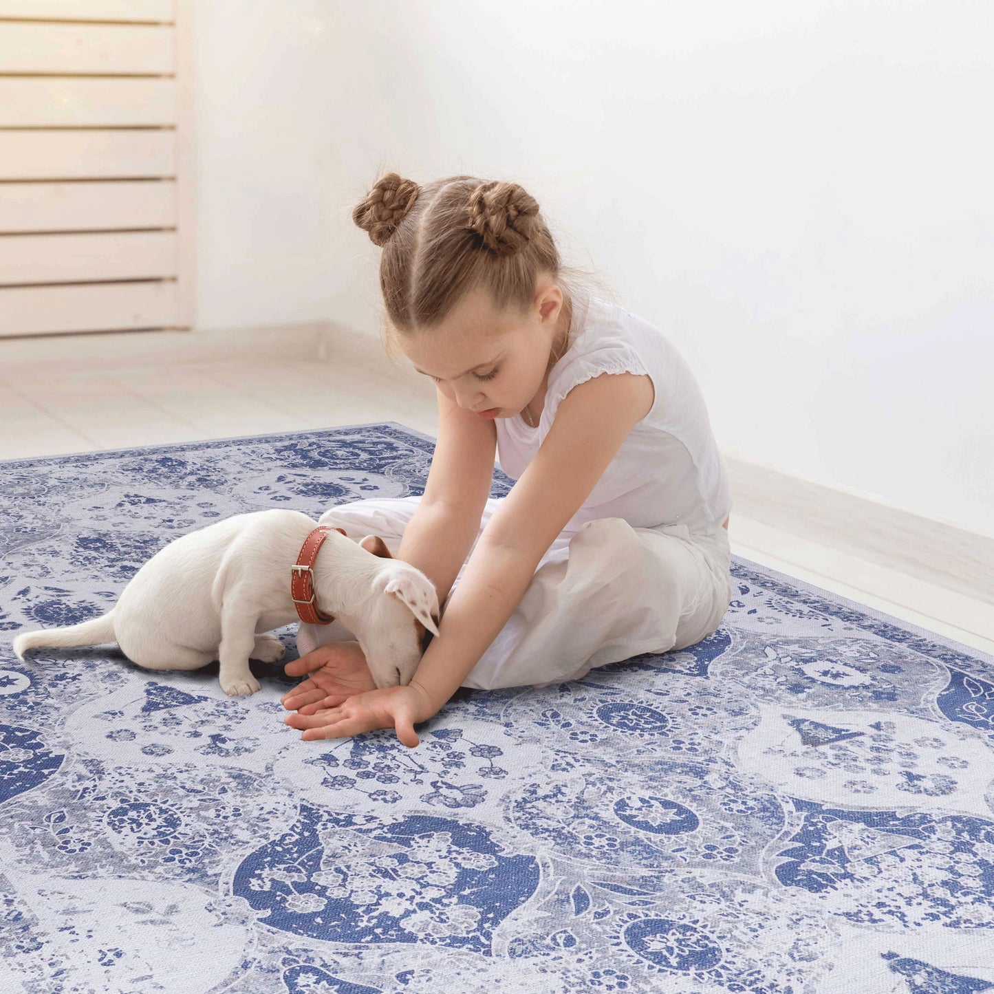 A young girl with braided hair sits on the Maval - Stunning Area Rug, gently playing with a small white puppy in a bright, minimal room with light-colored flooring.