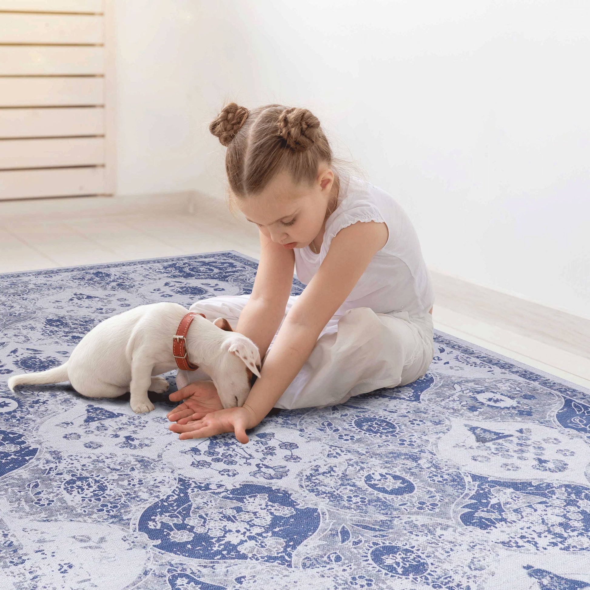 A young girl with braided hair sits on the Maval - Stunning Area Rug, gently playing with a small white puppy in a bright, minimal room with light-colored flooring.