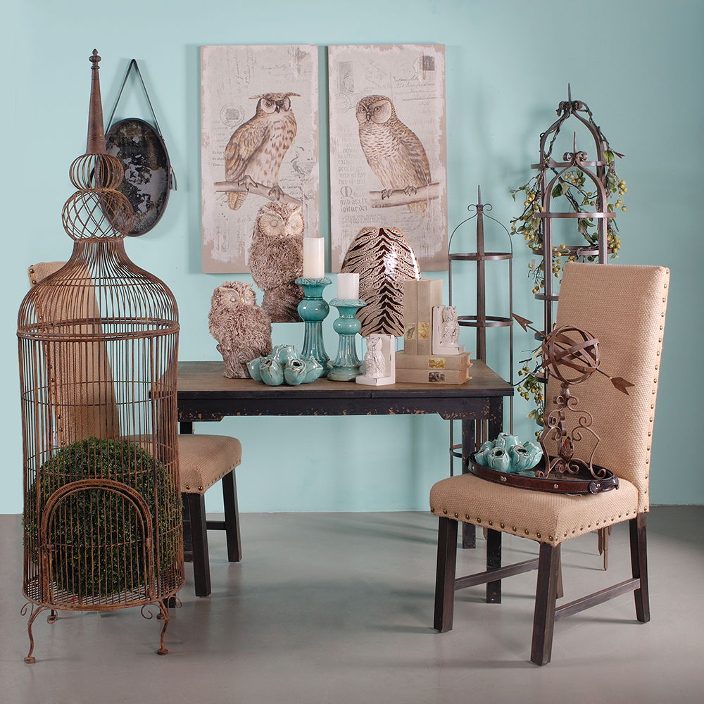 A room features a rustic table, two burlap chairs, decorative plants, owl figurines, turquoise vases, a metal birdcage, and Lilith Owl Prints With Distressed Look wall art (set of 2) hanging on a turquoise wall.