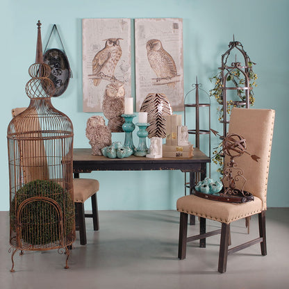 A room features a rustic table, two burlap chairs, decorative plants, owl figurines, turquoise vases, a metal birdcage, and Lilith Owl Prints With Distressed Look wall art (set of 2) hanging on a turquoise wall.