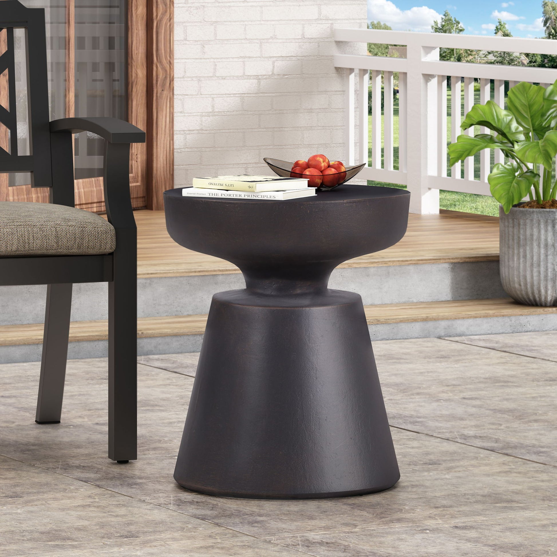 The Outdoor Lightweight Concrete End Table With Half Sphere Top - Copper features a modern dark brown finish and round top, perfect for patios. It holds books and a bowl of red apples next to a plant and chair.