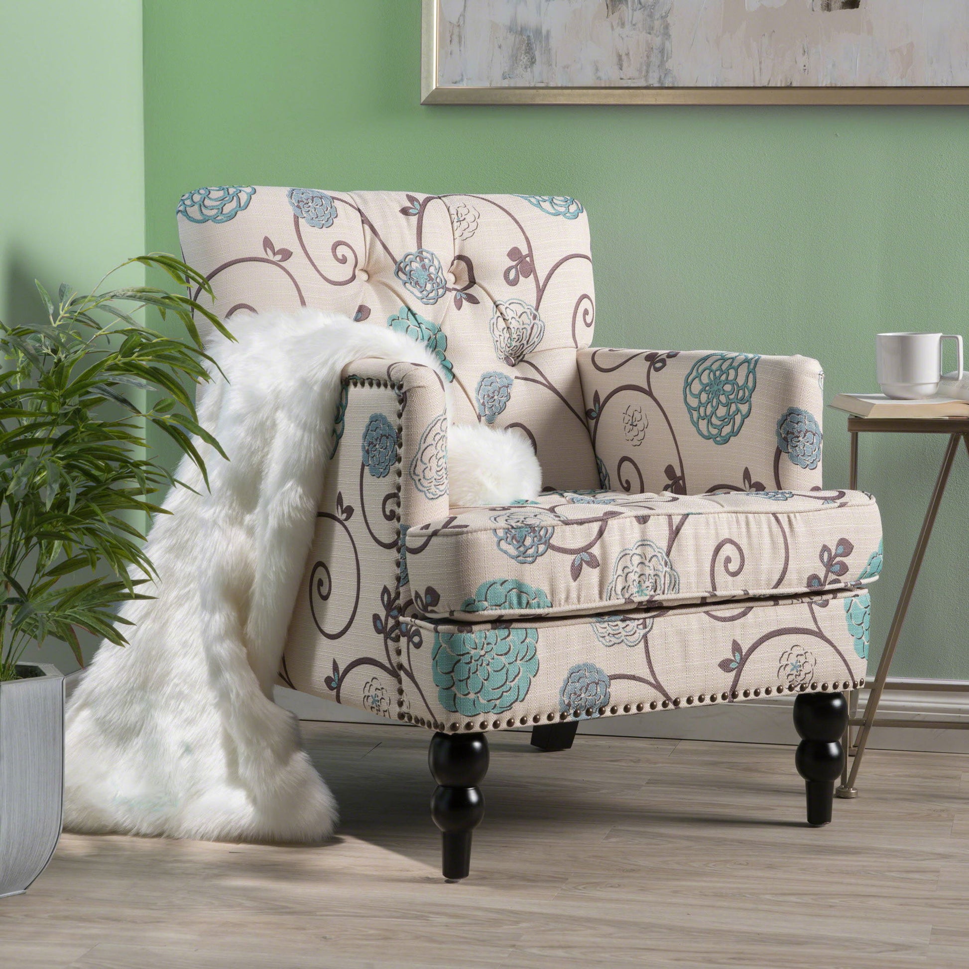The Vitality Club Chair, featuring button tufted diamond stitching and turned legs, sits on a wood floor near a small table and potted plant. A white faux fur throw drapes over the chair’s arm. The walls are painted light green.