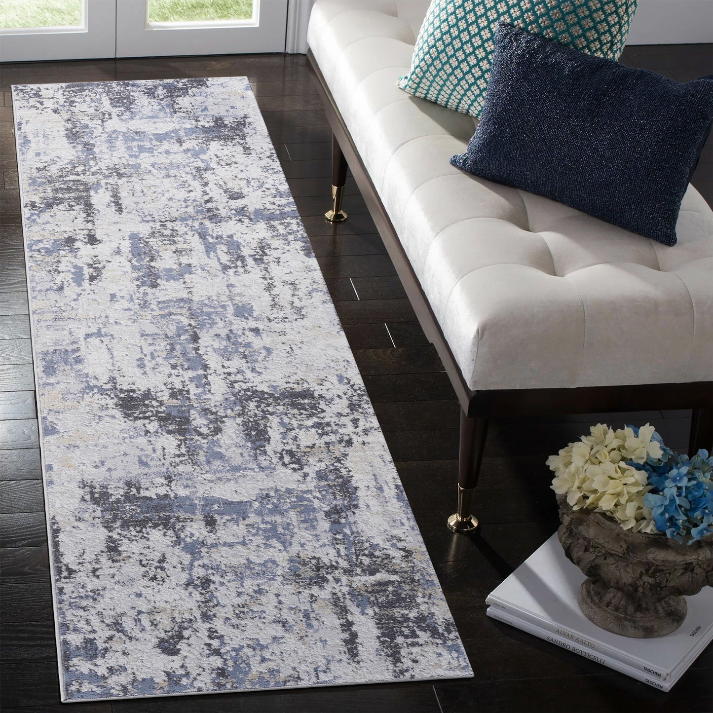 A modern hallway showcases the Payas - Abstract Area Rug in blue and gray tones on dark wood floors, paired with a white upholstered bench, blue accent pillows, and a decorative floral vase.