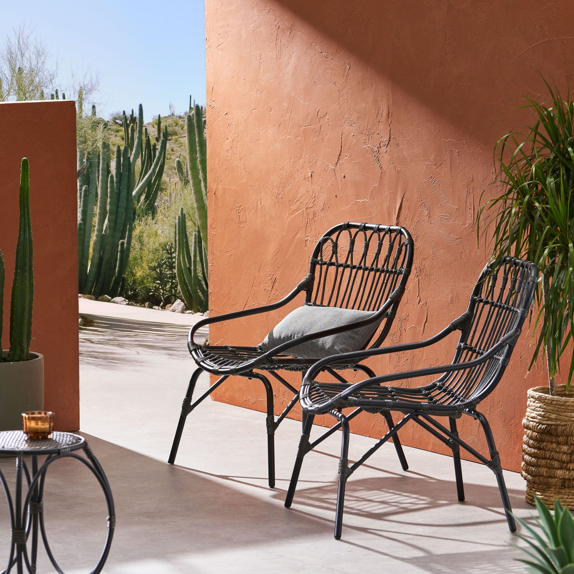 Two Monsoon Outdoor Accent Chairs with open weave design and gray cushions sit on a sunny patio by a terracotta wall, surrounded by potted plants and cacti. A small round table with a candle is in the foreground.