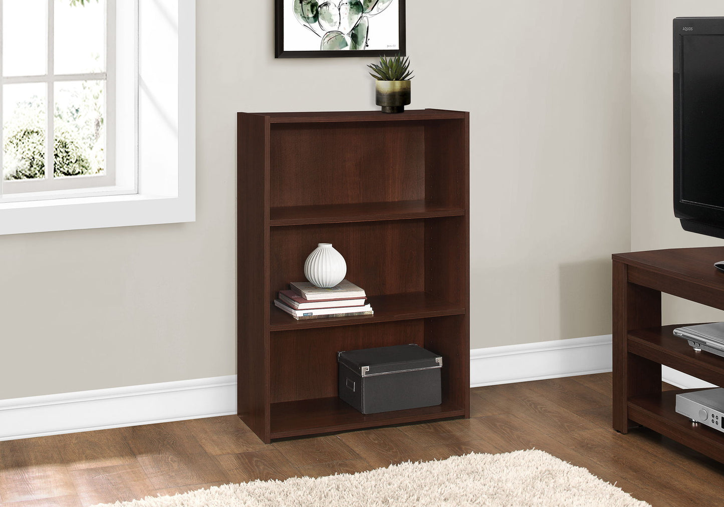 A 4-tier transitional bookcase holds books, a vase, a plant, and a storage box. Placed by a window against a light wall on hardwood flooring with a beige rug, this bookshelf is ideal for office spaces.