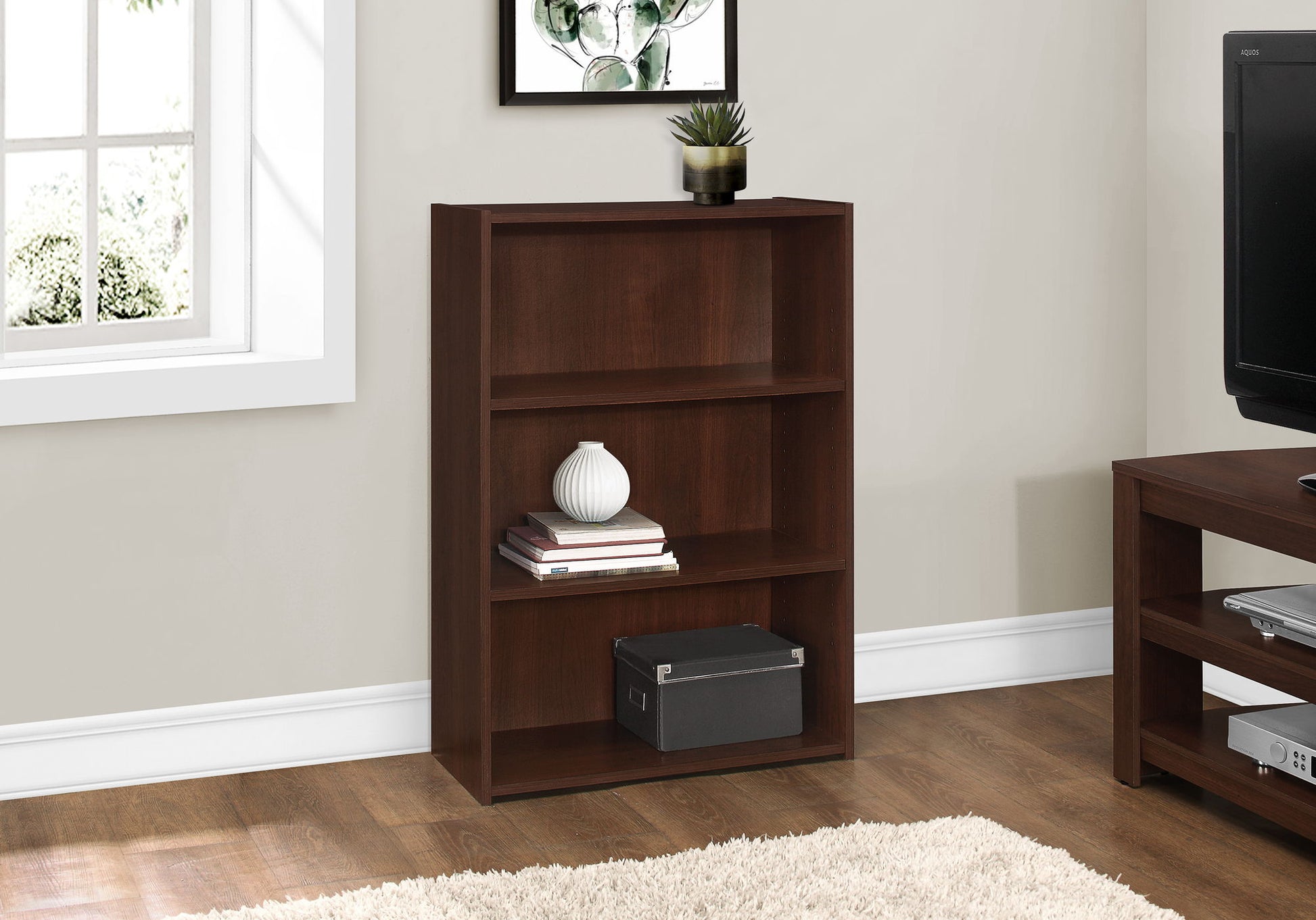 A 4-tier transitional bookcase holds books, a vase, a plant, and a storage box. Placed by a window against a light wall on hardwood flooring with a beige rug, this bookshelf is ideal for office spaces.