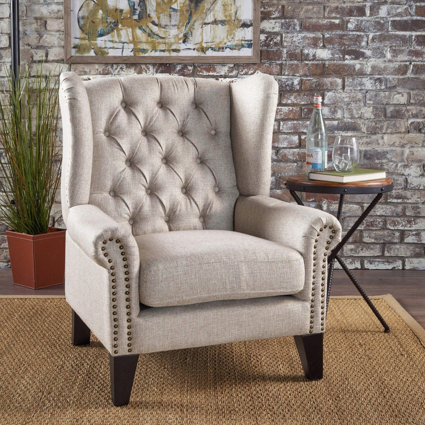 The Mid Century Modern Arm Reading Chair for Living Room, in beige with tufted wingback and studded trim, sits on a woven rug by a small table with a glass, bottle, books, and plant against a brick wall with abstract art.