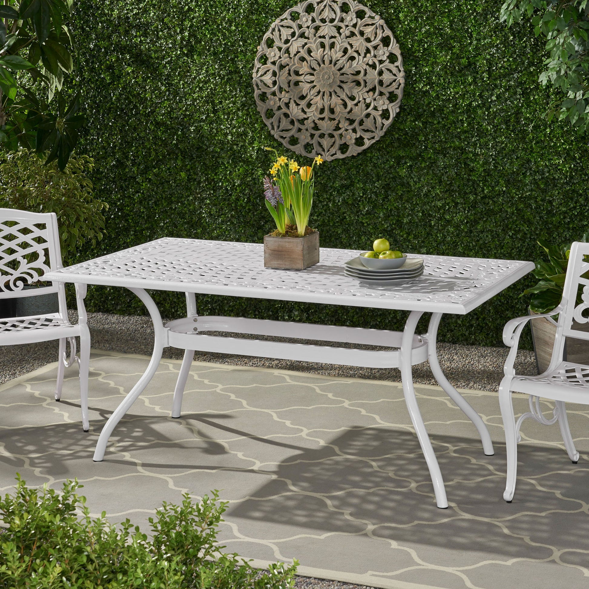 The Celestine Outdoor Cast Aluminum Dining Table with Floral Lattice Design and Umbrella Holder, paired with matching chairs, is set on a garden rug. It displays a potted yellow-flowered plant and green apples, backed by lush hedges and decorative wall art.